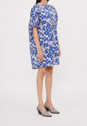 Femme portant une robe ample à fleurs bleues et blanches, longueur genoux, manches bouffantes, et des chaussures beiges à talons carrés, debout de profil.