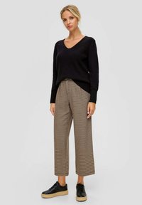 Vrouw in een zwarte V-hals trui, beige geruite wijde broek en zwarte sneakers met beige zolen, staande tegen een effen achtergrond.