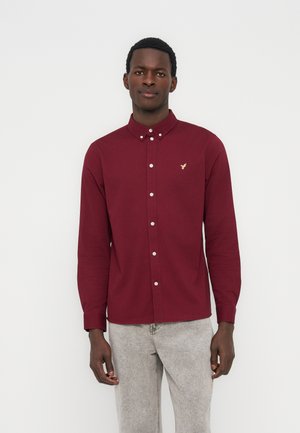 Άνδρας που φοράει πουκάμισο μπορντό με κουμπιά και μικρό λογότυπο στο στήθος και ανοιχτόχρωμο γκρι τζιν, στέκεται μπροστά σε λευκό φόντο.