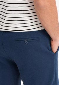 Marineblaue Hose aus strukturiertem Stoff mit einer Gesäßtasche und Knopfverschluss. Gestreiftes Oberteil darüber sichtbar, was einen Lagenlook kreiert.