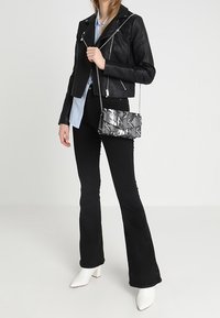 Chaqueta de motociclista de cuero negro con herrajes plateados, camisa azul claro, pantalones negros acampanados, bolso brillante de piel de serpiente plateado y botas blancas de tacón cuadrado.