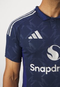 Námořnická modrá sportovní košile s texturovaným vzorem, má tři bílé pruhy na rameni, límec a logo na hrudi.