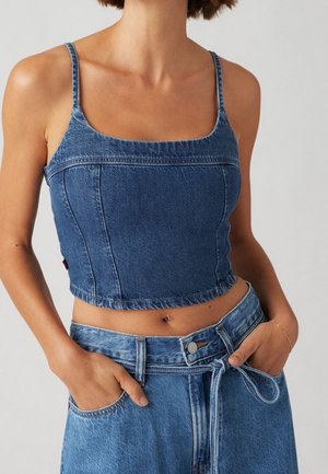 Nainen, jolla on sininen denimistä valmistettu lyhyt toppi ja korkeavyötäröiset denim-housut, kädet taskuissa yksiväristä taustaa vasten.