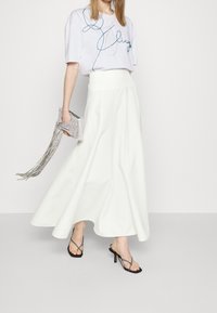 Falda larga blanca con un diseño fluido, combinada con una camiseta de color púrpura claro que presenta adornos azules, un bolso de mano plateado con flecos y sandalias negras de tacón.