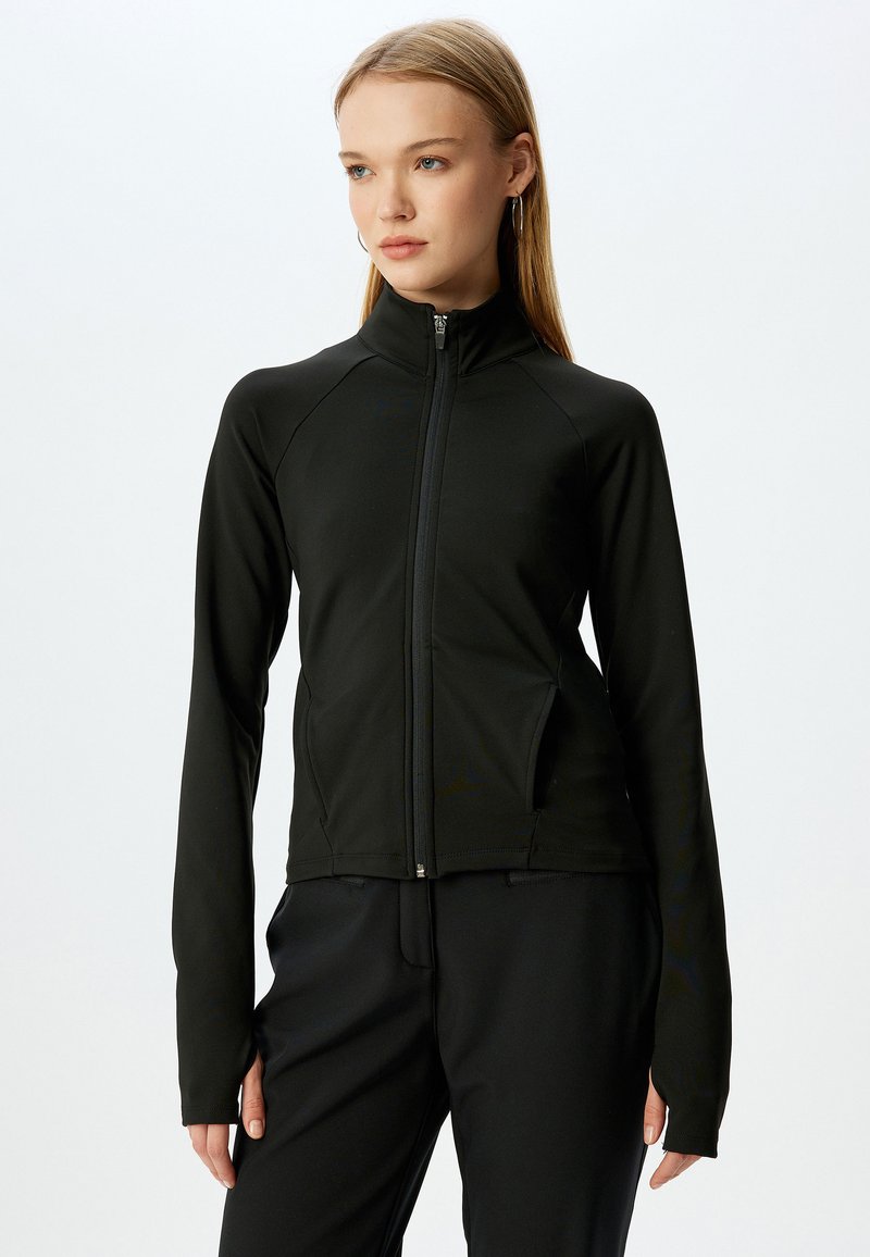 Chaqueta negra con cremallera y cuello alto, hecha de un tejido suave y elástico. Presenta mangas largas y bolsillos laterales. Diseño minimalista.