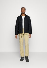 Sort quiltet jakke med lynlås foran, parret med beige bukser med en sidelomme og sorte accenter, samt solide sorte støvler.