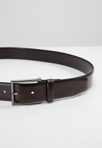 Ceinture en cuir marron avec une texture lisse, boucle en métal argenté, logo de la marque embossé et plusieurs trous d'ajustement.