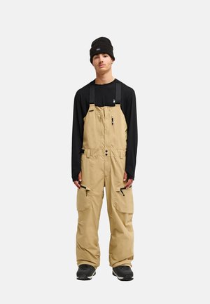 Junger Mann trägt beige Latzhose, schwarzes Langarmshirt, schwarze Mütze und schwarze Turnschuhe, steht vor einfachem Hintergrund.