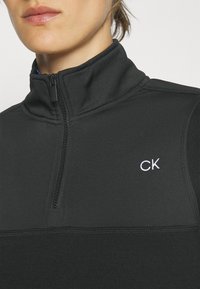 Veste zippée noire avec col montant, fabriquée en tissu doux. Présente un petit logo "CK" blanc et des accents texturés sur les épaules.
