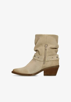 Beige suède enkellaars met een slouchy schacht, zijrits, decoratief genaaide riem met metalen oogjes en een houten blokhak.