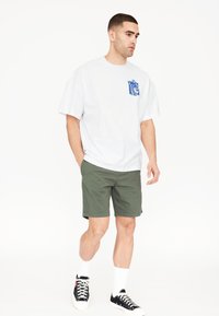 Camiseta blanca de gran tamaño con un bolsillo gráfico azul, combinada con pantalones cortos verdes y zapatillas negras, que presenta calcetines blancos y una postura casual.
