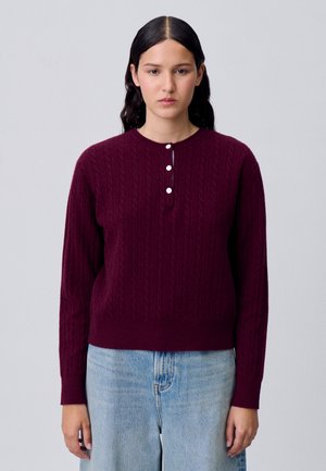 Pull en maille torsadée bordeaux avec un col rond, doté d'une patte de boutonnage et de poignets côtelés, associé à un jean en denim bleu clair.