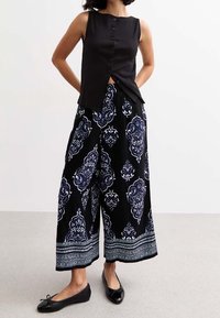 Černý bezrukávový top s knoflíky, spárovaný s širokými kalhotami s bílým a modrým zdobeným vzorem a dekorativním lemem. Na nohách černé baleríny.