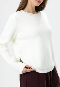 Pull blanc en matériau tricoté doux, avec un col rond côtelé, des boutons sur le côté et un ourlet incurvé pour une coupe décontractée.