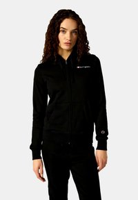 Schwarze Zip-Hoodie aus weichem Stoff, mit einer Fronttasche, gerippten Bündchen und einem kleinen Logo auf der Brust und dem Ärmel.