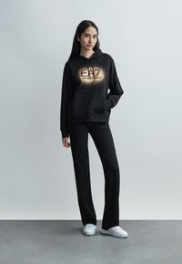 Μαύρο φούτερ με κουκούλα, μπροστινή τσέπη και ανάγλυφο χρυσό λογότυπο, σε συνδυασμό με μαύρο παντελόνι και λευκά αθλητικά παπούτσια, σε φόντο ανοιχτό γκρι.