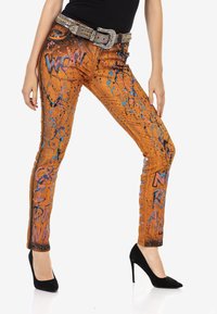 Jeans en denim orange avec des motifs de éclaboussures bleues et roses, présentant des coutures visibles, des poches latérales et une ceinture embellie. Chaussures à talons noirs.