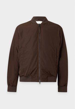 Homme portant une veste marron zippée, une chemise verte, un jean bleu évasé et des baskets marron, debout devant un fond blanc.