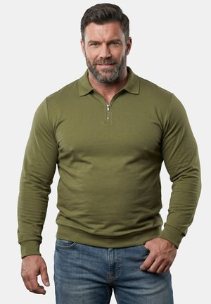 Skæggede mand med kort mørkt hår iført en olivengrøn langærmet skjorte med lynlås foran og blå jeans, stående med den ene hånd i lommen.