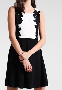 Robe noire et blanche avec un corsage ajusté orné d'appliqués en dentelle florale, un col rond et une jupe évasée. Texture de tissu lisse.