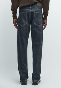 Tmavě džínové kalhoty s volnějším střihem, vybavené dvěma zadními kapsami a prošitými švy. Nošeny s hnědým texturovaným svetrem a černými botami.