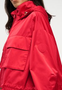 Γυναίκα με φωτεινό κόκκινο μπουφάν με κουκούλα, μεγάλες εμπρόσθιες τσέπες και στρογγυλή λογότυπο ραμμένο στο μανίκι.