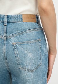 Personne portant un jean en denim bleu clair avec des motifs abstraits blancs et une étiquette en cuir marron de la marque Samsøe Samsøe à la taille arrière.