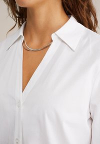Camicia bianca con colletto a punta e design a V. Presenta una leggera texture e un tessuto traslucido. Collana doppia in argento.