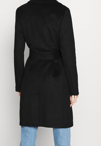 Zwarte wollen jas met een ceintuur in de taille, met een knoopkraag en lange mouwen. De stof lijkt zacht met een gladde textuur.