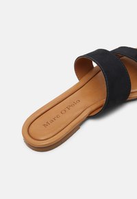 Infradito con cuscinetto di colore beige, dotate di una ampia fascia in suede nera e con il logo "Marc O'Polo" stampato sulla soletta, il tutto su uno sfondo bianco.