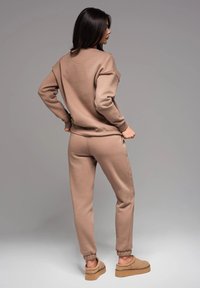 Brauner Baumwollpullover und passende Sweatpants mit elastischen Bündchen. Das Modell steht mit dem Rücken zur Kamera auf einem grauen Hintergrund und trägt beigen Slipper.