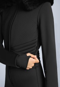 Osoba w czarnej pikowanej kurtce z futrzanym kołnierzem, trzymająca zamek w okolicy talii, na jednolitym jasnym tle.