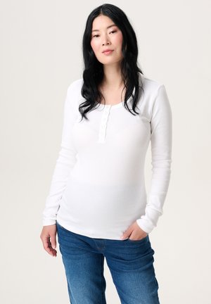 Frau mit langen schwarzen Haaren, die ein weißes, langärmeliges Hemd mit Knöpfen und blaue Jeans trägt, steht mit einer Hand in der Tasche.