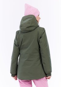 Grüne wasserfeste Jacke mit Kapuze, mit elastischen Bündchen und geradem Schnitt. Kombiniert mit hellrosa Hosen und einer pinken Mütze.