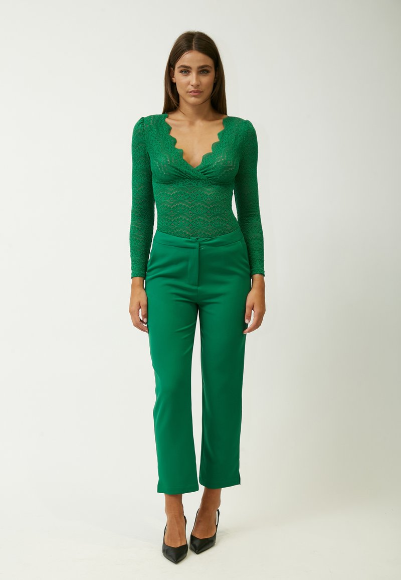 Groene kanten bodysuit met een golvende halslijn, gecombineerd met op maat gemaakte groene broeken. Zwarte hoge hakken maken de outfit compleet. Eenvoudige achtergrond.