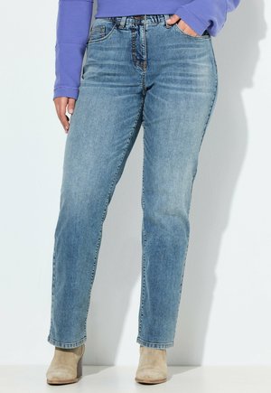 Osoba ubrana w jasnoniebieskie jeansy o prostych nogawkach, beżowe botki za kostkę i fioletowy długi rękaw, z jedną ręką w kieszeni.