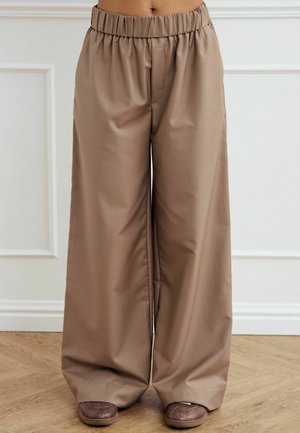 Beige weite Hosen mit elastischem Bund, mit glatter Textur und lockerem Sitz, kombiniert mit braunen Hausschuhen auf einem Holzboden.