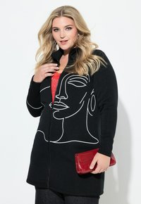 Czarna kurtka na zamek z białym rysunkiem twarzy, wykonana z miękkiego materiału. Modelka trzyma w jednej ręce czerwony błyszczący kopertówkę.