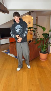 Šedé kalhoty s širokými nohavicemi, černá mikina s kapucí s motivem vlka a bílými akcenty. Bílé tenisky. Vnitřní prostředí s zelenou rostlinou a dřevěnou podlahou.