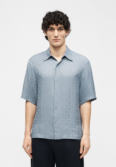 Άνδρας με σγουρά μαλλιά που φοράει κοντομάνικο πουκάμισο ανοιχτού μπλε με μοτίβο, στέκεται μπροστά σε απλό λευκό φόντο.