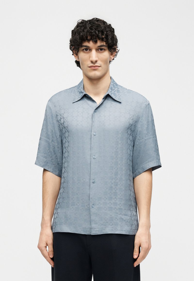 Man with curly hair wearing short-sleeve light blue patterned button-up shirt, standing against plain white background.