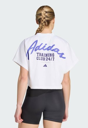 Kvinde med flettet hår iført hvid kort Adidas-shirt med teksten "Training Club 24/7" og sorte træningsshorts, vendt væk.