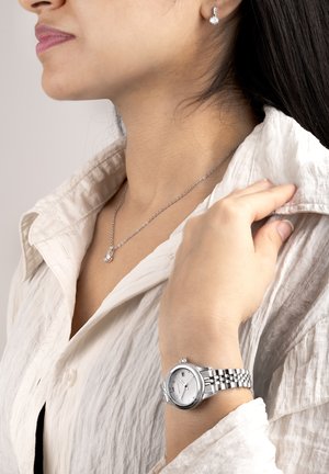 Silberne Uhr mit rundem weißen Zifferblatt, metallischem Armband und Datumsanzeige. Kombiniert mit einer schlichten silbernen Halskette und Ohrringen. Zerknittertes weißes Hemd.