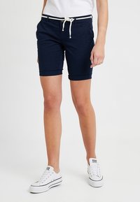 Personne portant un short bleu marine avec une ceinture en corde blanche, un T-shirt blanc et des baskets basses blanches, debout sur un fond blanc.
