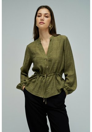 Femme portant un chemisier à manches longues vert olive avec taille ajustable par cordon, pantalon noir et boucles d'oreilles dorées, debout les mains dans les poches devant un fond uni.
