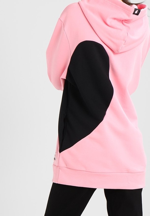 Personne portant un sweat à capuche rose ample avec un grand patch noir en forme de cœur dans le dos, associé à un pantalon noir.