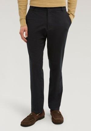 Mann trägt schwarze, gerade geschnittene Hosen, braune Wildleder-Loafers und ein beiges, strukturiertes Langarmshirt, steht mit einer Hand in der Tasche.