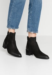 Botines de ante negro con punta afilada y pequeño tacón cuadrado. Cuentan con cremalleras laterales y un diseño elegante para un uso fácil.