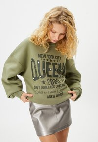 Sudadera verde oliva de corte suelto, con texto impreso en negro y estrellas decorativas, combinada con una mini falda plateada brillante.