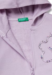 Sudadera de color púrpura claro con cremallera, forro interior texturizado, diseño floral bordado y una etiqueta verde que dice "UNITED COLORS OF BENETTON."
